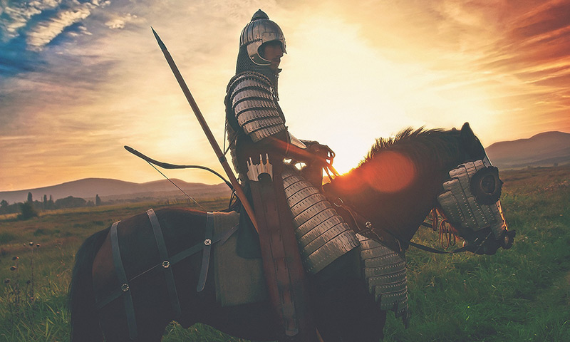 Photo of a knight sitting on on a horse in a field at sunset.